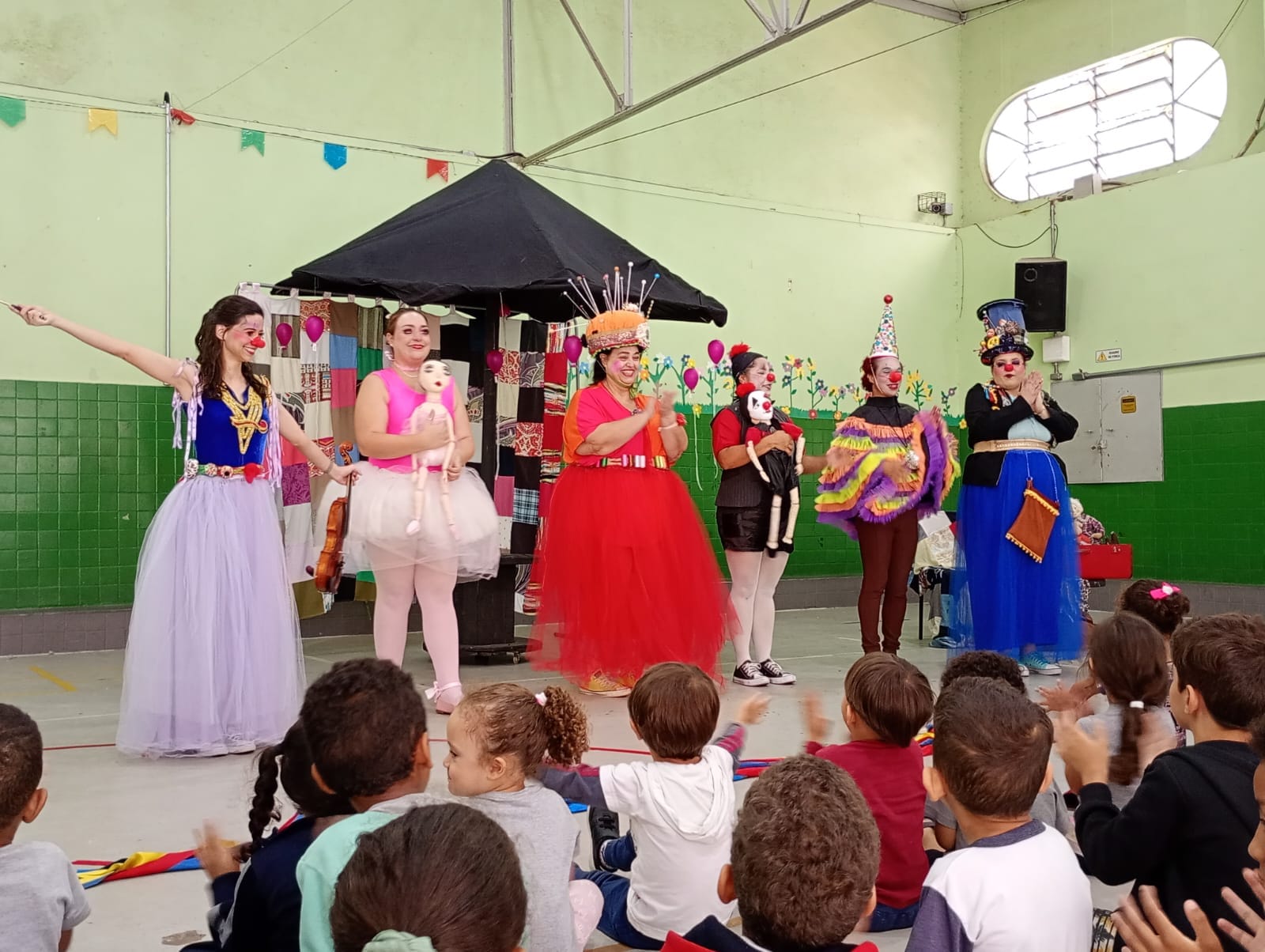 Apresentação teatral da Cia Casulo para crianças. Seis artistas caracterizados em personagens e figurinos coloridos estão em cena diante de um grupo de crianças sentadas, que acompanha a apresentação. Ao fundo, há um cenário cênico com tendas, tecidos e elementos decorativos.
